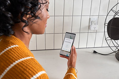 A woman holds a phone and checks the smart plug in the app. In front is an old-style grille fan and a smart plug inserted into a wall outlet   